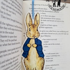 Può includere: Un segnalibro in feltro con Peter Rabbit dal libro per bambini di Beatrix Potter. Il segnalibro è blu e bianco con un retro in feltro marrone. Il segnalibro è fatto a mano e ha il testo "Handmade".