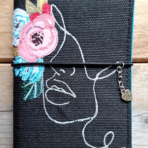 May include: A black fabric notebook cover featuring a white line art profile of a face and embroidered floral accents in pink, blue, and gold. A black elastic band secures the cover, adorned with a small silver heart charm labeled Made with Love.