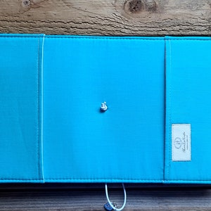 Peut inclure: Une couverture de carnet en tissu bleu turquoise avec une fermeture &agrave; bouton blanc et une petite &eacute;tiquette blanche avec le texte "Handmade" sur le c&ocirc;t&eacute; droit.