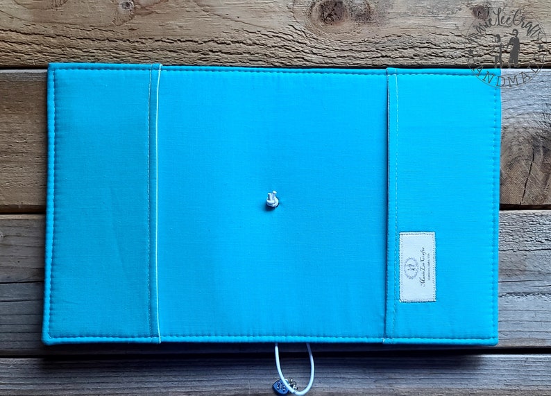 Peut inclure: Une couverture de carnet en tissu bleu turquoise avec une fermeture &agrave; bouton blanc et une petite &eacute;tiquette blanche avec le texte "Handmade" sur le c&ocirc;t&eacute; droit.