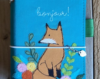 Könnte beinhalten: Ein türkisfarbener Notizbuchumschlag aus Stoff mit einer Stickerei eines braunen Fuchses, der zwischen bunten Blumen und grünen Blättern sitzt. Das Wort bonjour! ist in weißem Garn über dem Fuchs gestickt. Ein weißes Gummiband hält einen Metallanhänger.