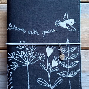 Peut inclure: Couverture de carnet en lin noir avec broderie blanche de fleurs, un oiseau et le texte "Bloom with grace". Un charme argent&eacute; avec le texte "Made with Love" pend du carnet.