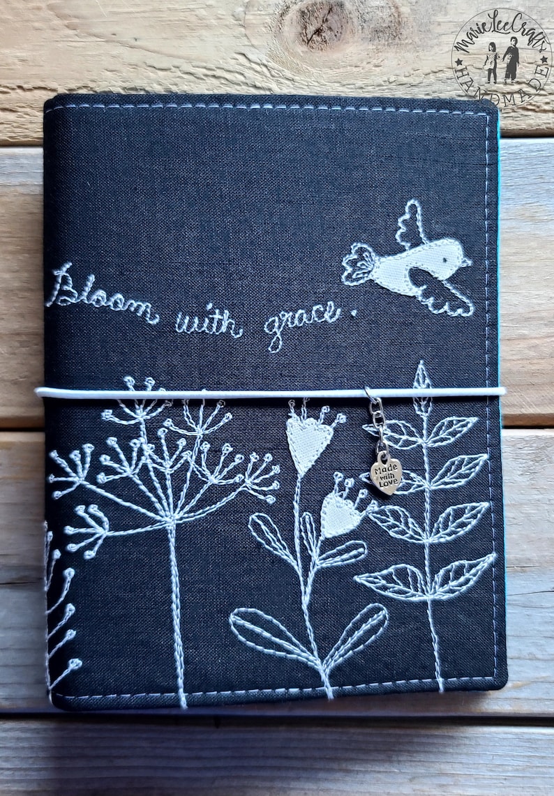 Peut inclure: Couverture de carnet en lin noir avec broderie blanche de fleurs, un oiseau et le texte "Bloom with grace". Un charme argent&eacute; avec le texte "Made with Love" pend du carnet.