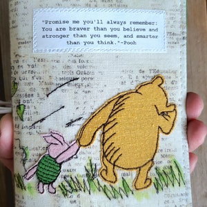 Può includere: Un ricamo con Winnie the Pooh e Maialino su uno sfondo di tessuto bianco con una citazione di Winnie the Pooh: "Promettimi che ti ricorderai sempre: Sei più coraggioso di quanto pensi e più forte di quanto sembri, e più intelligente di quanto pensi." - Winnie the Pooh