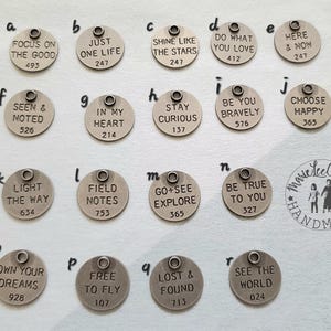 May include: A set of 16 silver metal charms with inspirational quotes and numbers. The charms are round and have a hole at the top for hanging. The quotes include "Focus on the good", "Just one life", "Shine like the stars", "Do what you love", "Here & now", "Seen & noted", "In my heart", "Stay curious", "Be you bravely", "Choose happy", "Light the way", "Field notes", "Go + see explore", "Be true to you", "Own your dreams", "Free to fly", "Lost & found", and "See the world".
