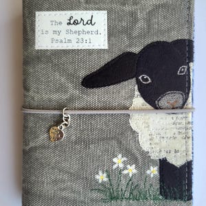 May include: A gray fabric book cover featuring an embroidered black sheep, white flowers, and a white rectangular patch with the text "The Lord is my Shepherd. Psalm 23:1". A gray elastic band holds a silver heart charm engraved with "made with love".