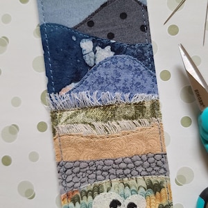 Puede incluir: Marcapáginas de tela hecho a mano con un diseño de paisaje. El marcapáginas presenta capas de telas azules, grises y beige, con un diseño de ovejas en la parte inferior. La parte superior tiene el texto "Marie Lee Crafts Handmade".