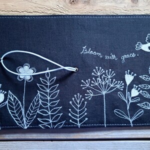 Peut inclure: Une couverture de carnet en lin noir avec des broderies blanches de fleurs, de feuilles et d'un oiseau. La couverture est dot&eacute;e d'une sangle blanche avec un fermoir m&eacute;tallique et le texte "Bloom with grace." est brod&eacute; sur la couverture.