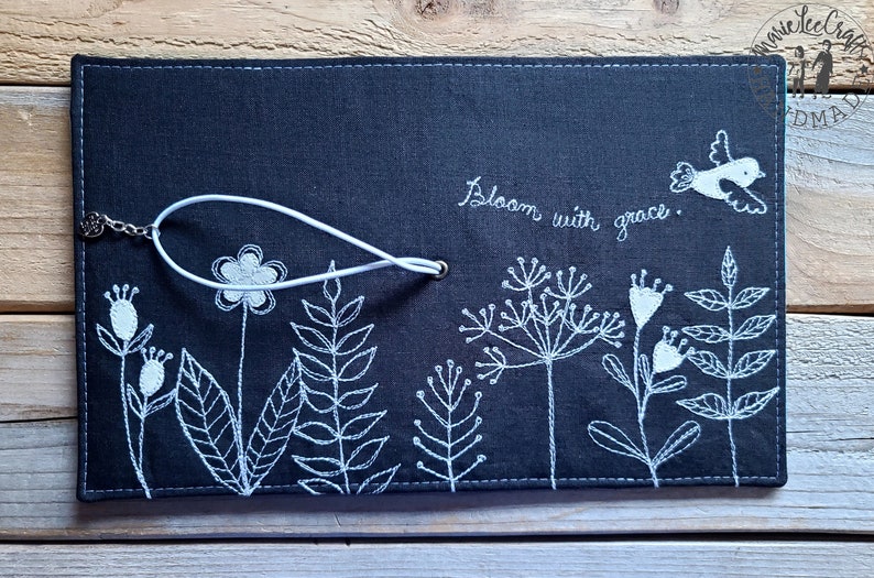 Peut inclure: Une couverture de carnet en lin noir avec des broderies blanches de fleurs, de feuilles et d'un oiseau. La couverture est dot&eacute;e d'une sangle blanche avec un fermoir m&eacute;tallique et le texte "Bloom with grace." est brod&eacute; sur la couverture.