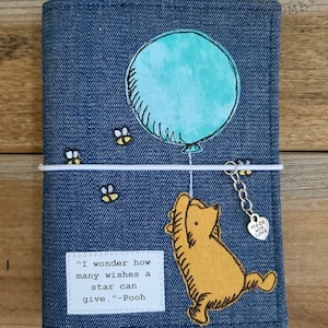 May include: A blue denim notebook cover with a Winnie the Pooh illustration of Pooh holding a blue balloon. The quote "I wonder how many wishes a star can give. - Pooh" is printed on the cover. A silver charm with the words "Made with Love" is attached to the notebook.