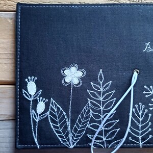 Peut inclure: Un carnet en tissu noir avec des broderies blanches de fleurs et de feuilles. Le carnet est dot&eacute; d'un cordon blanc avec un charme en forme de c&oelig;ur.