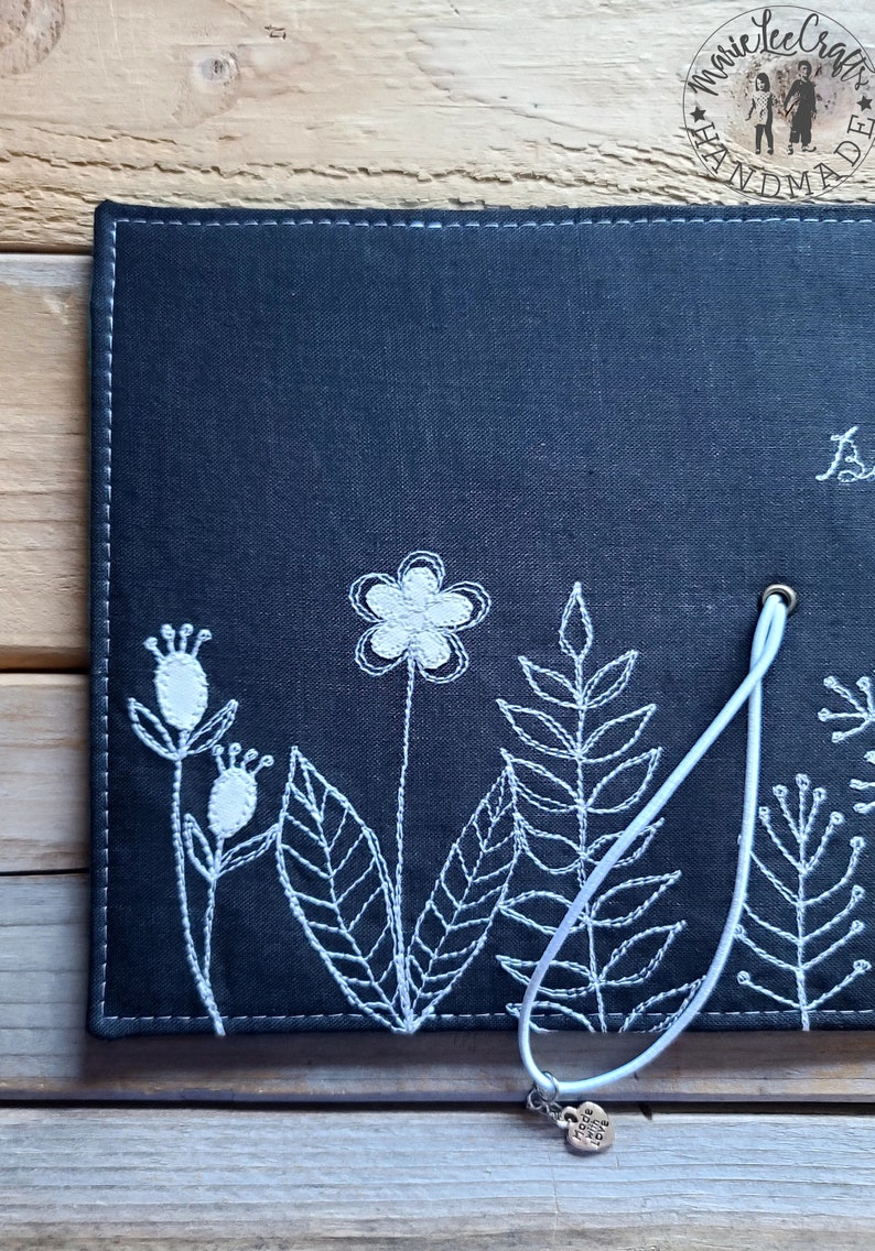 Peut inclure: Un carnet en tissu noir avec des broderies blanches de fleurs et de feuilles. Le carnet est dot&eacute; d'un cordon blanc avec un charme en forme de c&oelig;ur.