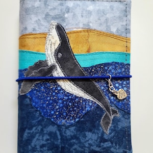 Puede incluir: Una funda de tela para cuaderno con una ballena negra y blanca aplicada nadando en un mar azul. La funda incluye bandas horizontales de tela gris, amarilla y turquesa, un cierre elástico azul y un dije de corazón plateado con la inscripción Made with Love.