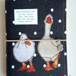 May include: A black fabric notebook cover with white polka dots featuring two geese illustrations and a white rectangular label with the text: Good friends lift you up, speak life, and walk with you through every season. A tan elastic band holds a heart charm.