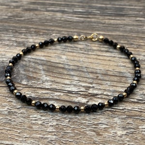 May include: Delicate black beaded bracelet with gold-colored accents. The bracelet features small, faceted black beads interspersed with gold-colored beads and a gold-colored clasp. The jewelry is displayed on a wooden surface.