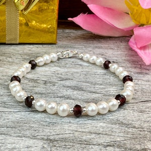 May include: A white pearl bracelet with small, dark red gemstone accents. The bracelet is on a wooden surface with a pink flower and a gold gift box in the background.