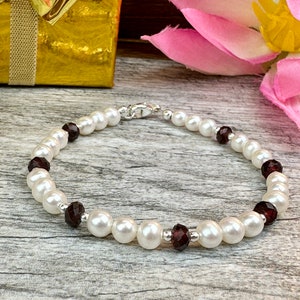 May include: A white pearl bracelet with dark red gemstone accents. The bracelet is on a wooden surface.