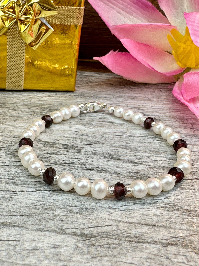 May include: A white pearl bracelet with dark red gemstone accents. The bracelet is on a wooden surface.