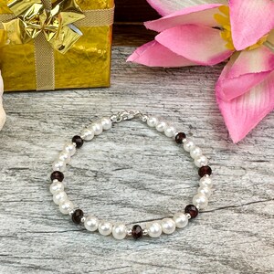 May include: A white pearl bracelet with dark red gemstone accents. The bracelet is on a wooden surface.