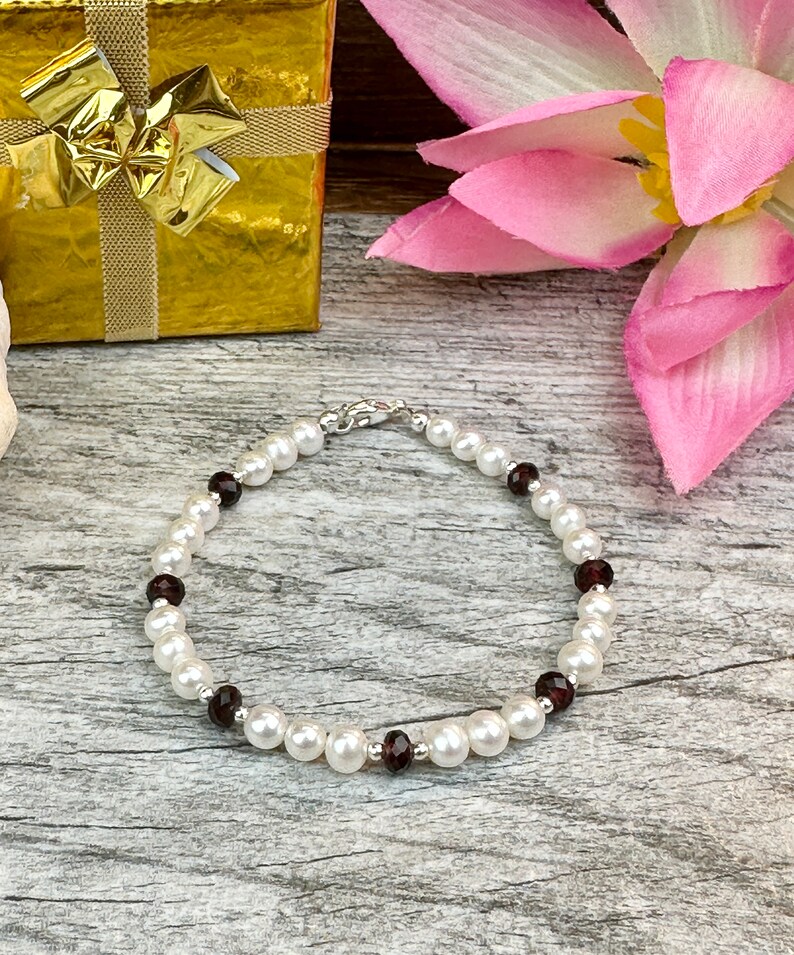 May include: A white pearl bracelet with dark red gemstone accents. The bracelet is on a wooden surface.