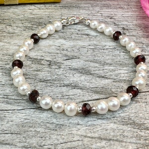 May include: A white pearl and garnet bracelet with a silver clasp. The bracelet is on a wooden surface.