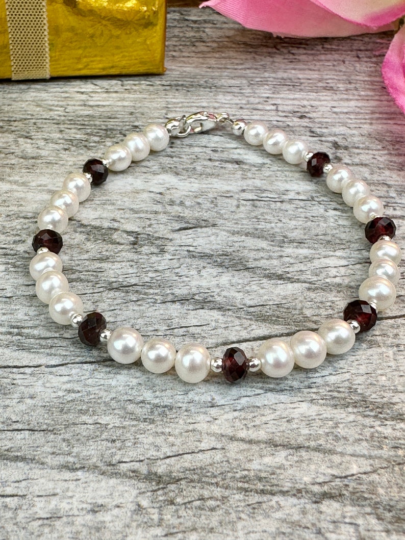 May include: A white pearl and garnet bracelet with a silver clasp. The bracelet is on a wooden surface.