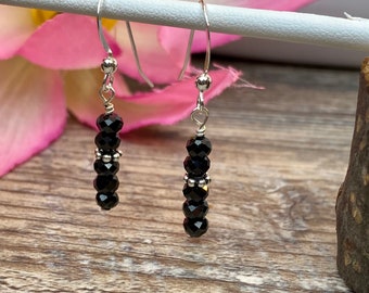 May include: A pair of silver dangle earrings with black crystal beads. The earrings are hanging from a white surface.