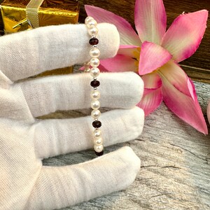 May include: A white pearl bracelet with small red gemstone accents. The bracelet is on a white surface.