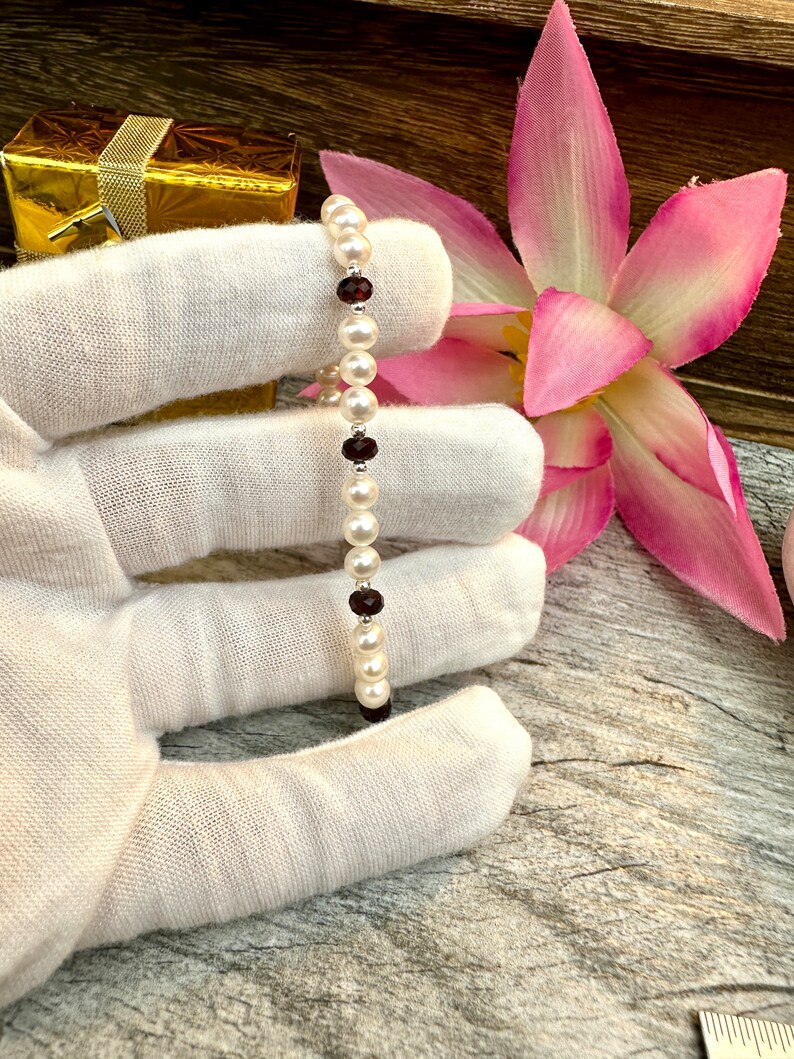 May include: A white pearl bracelet with small red gemstone accents. The bracelet is on a white surface.