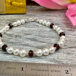 May include: A white pearl bracelet with dark red gemstone accents. The bracelet is approximately 7 inches long and is measured on a ruler.