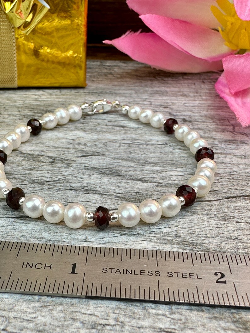 May include: A white pearl bracelet with dark red gemstone accents. The bracelet is approximately 7 inches long and is measured on a ruler.