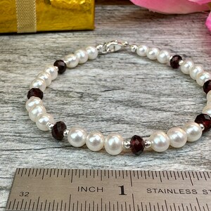 May include: A white pearl bracelet with dark red gemstone accents. The bracelet is approximately 1 inch in length and is measured against a ruler.