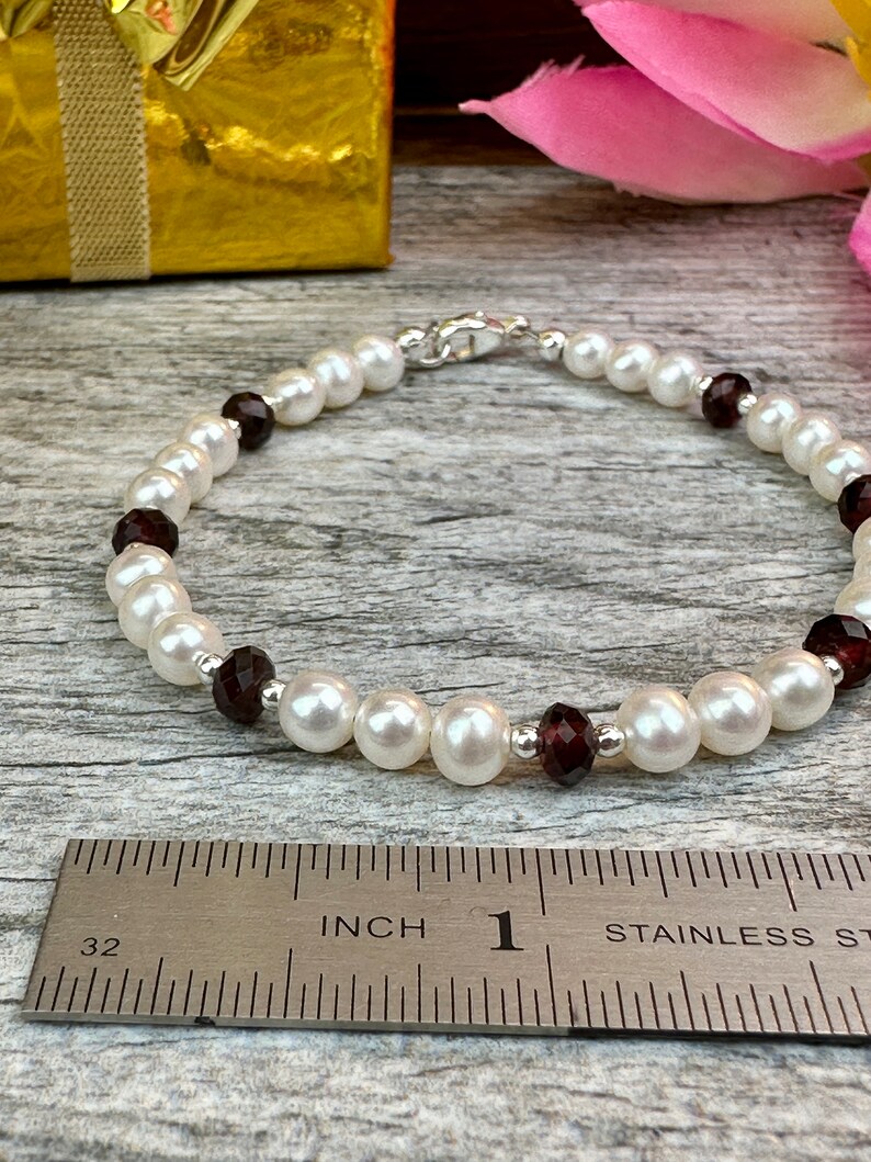 May include: A white pearl bracelet with dark red gemstone accents. The bracelet is approximately 1 inch in length and is measured against a ruler.