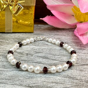 May include: A white pearl bracelet with dark red gemstone accents. The bracelet is on a wooden surface with a gold gift box and a pink flower in the background.