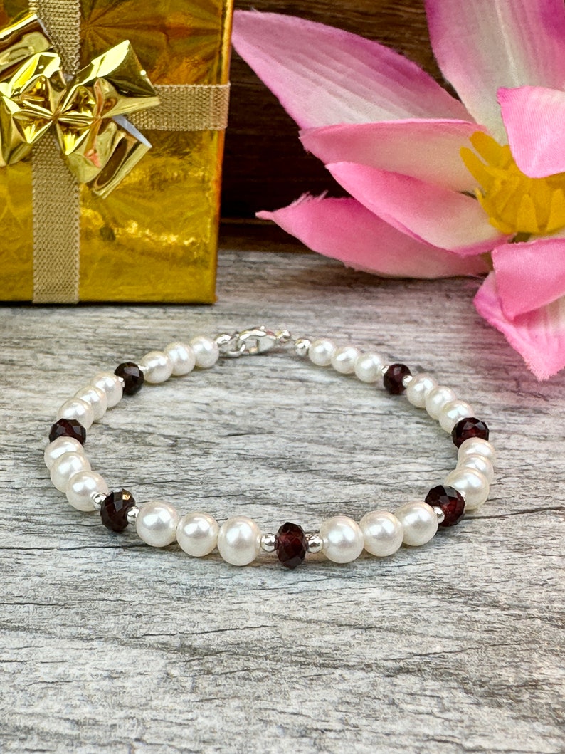 May include: A white pearl bracelet with dark red gemstone accents. The bracelet is on a wooden surface with a gold gift box and a pink flower in the background.