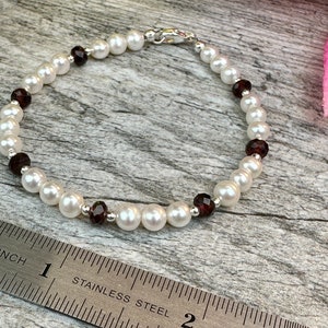 May include: A white pearl and garnet bracelet with a silver clasp. The bracelet is measured on a ruler, showing it is about 7 inches long.