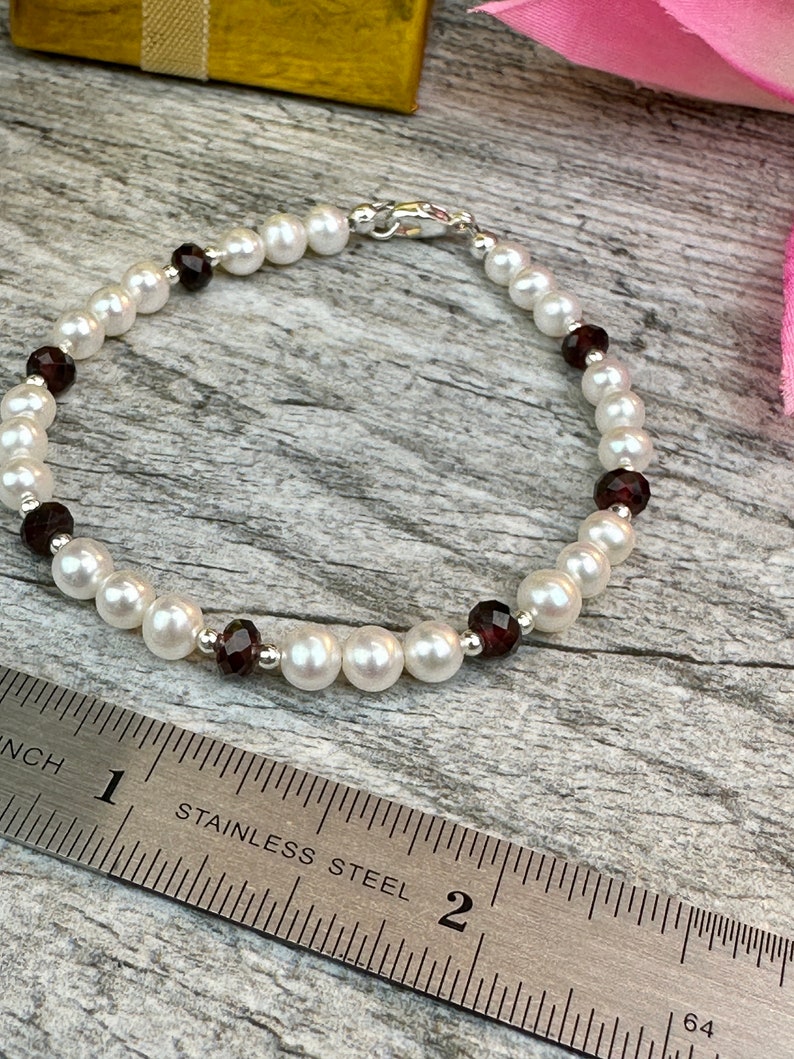 May include: A white pearl and garnet bracelet with a silver clasp. The bracelet is measured on a ruler, showing it is about 7 inches long.