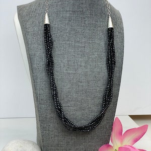 Puede incluir: Un collar de múltiples hilos hecho de cuentas negras con tapas de extremo plateadas. El collar se muestra en un maniquí gris.