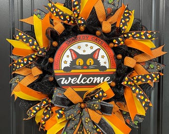 May include: A Halloween wreath featuring a black mesh base with orange, yellow, and black patterned ribbons. A central circular sign displays a black cat with yellow eyes and the text "SCAREDY CATS" and "welcome" in white and orange lettering.