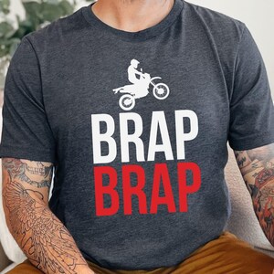 May include: A dark gray t-shirt with a white silhouette of a person riding a dirt bike. The text "BRAP BRAP" is printed on the shirt, with the top word in white and the bottom word in red.