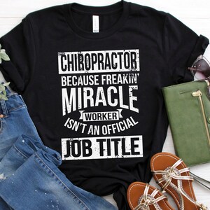 May include: Black t-shirt with white text that reads "Chiropractor because freakin' miracle worker isn't an official job title". The shirt is laid out on a white wooden surface with blue jeans, sandals, and a green wallet.