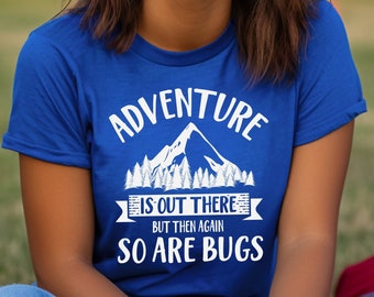 May include: A royal blue short-sleeve t-shirt featuring a white graphic print of a mountain range and pine trees. The text reads ADVENTURE IS OUT THERE BUT THEN AGAIN SO ARE BUGS in a bold, white, sans-serif font centered on the chest area.