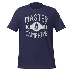 May include: A navy blue short-sleeve t-shirt featuring a white distressed graphic on the chest. The design includes a central campfire icon inside a circle, surrounded by the text "MASTER OF THE CAMPFIRE" in a bold, capitalized, sans-serif font.