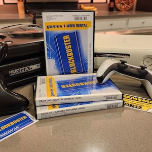 May include: A collection of video game consoles and games from the 1990s and 2000s, including a Sega Genesis, an Xbox 360, and a stack of Blockbuster video game rentals. The games are Mortal Kombat (2011) for Xbox 360 and Batman Returns (1992) for Sega CD.