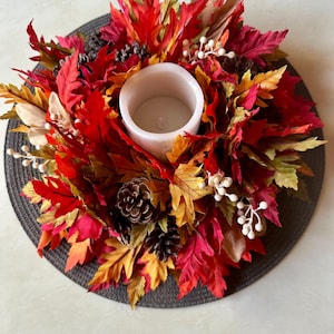 K&ouml;nnte beinhalten: Ein herbstliches Kerzenhalter-Arrangement mit einer wei&szlig;en Kerze, umgeben von leuchtend roten, orangefarbenen und gelben Bl&auml;ttern. Die Dekoration umfasst Tannenzapfen und wei&szlig;e Beeren, alles auf einer dunkelgrauen runden Basis.