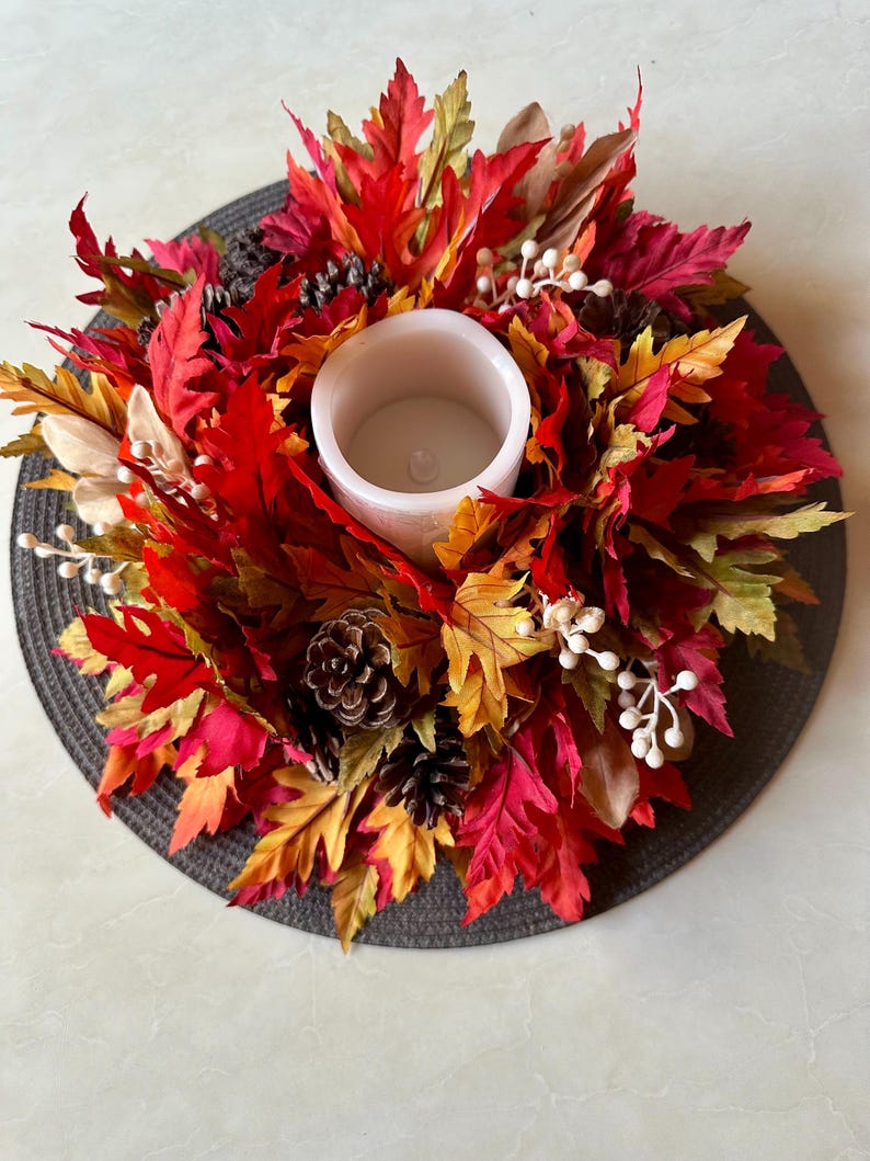 K&ouml;nnte beinhalten: Ein herbstliches Kerzenhalter-Arrangement mit einer wei&szlig;en Kerze, umgeben von leuchtend roten, orangefarbenen und gelben Bl&auml;ttern. Die Dekoration umfasst Tannenzapfen und wei&szlig;e Beeren, alles auf einer dunkelgrauen runden Basis.
