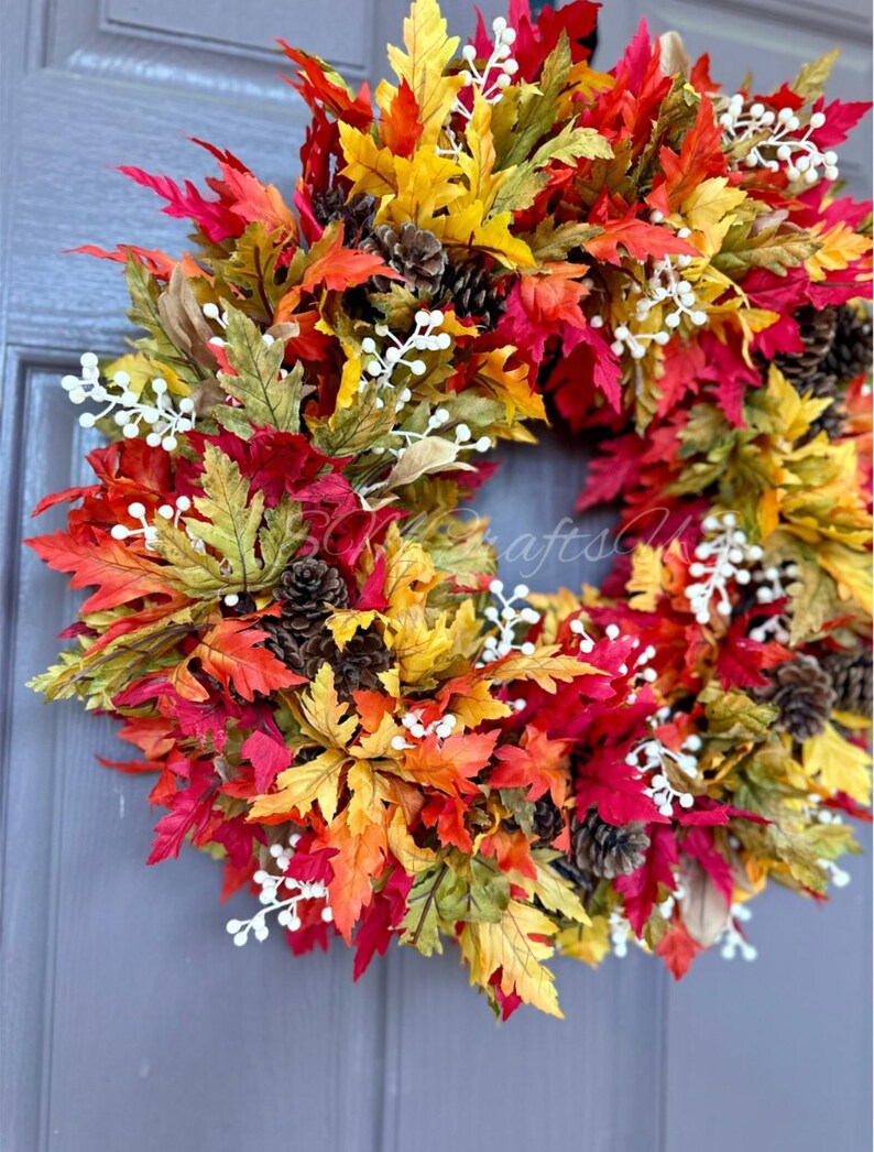 K&ouml;nnte beinhalten: Ein lebendiger Herbstkranz mit bunten Ahornbl&auml;ttern in Rot-, Orange- und Gelbt&ouml;nen. Der Kranz ist mit kleinen Tannenzapfen und wei&szlig;en Beerenzweigen geschm&uuml;ckt und schafft eine festliche saisonale Dekoration.