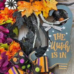 May include: A Halloween wreath with a black witch's boot sign that says "The Witch Is In". The wreath is decorated with orange, purple, and black fall leaves, a black and white pumpkin, and a black and orange striped bow with green and purple polka dots.