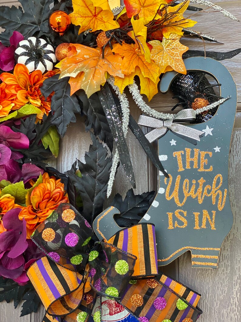 May include: A Halloween wreath with a black witch's boot sign that says "The Witch Is In". The wreath is decorated with orange, purple, and black fall leaves, a black and white pumpkin, and a black and orange striped bow with green and purple polka dots.