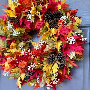 K&ouml;nnte beinhalten: Ein farbenfroher Herbstkranz mit roten, gelben und gr&uuml;nen Bl&auml;ttern. Der Kranz ist mit Tannenzapfen und kleinen wei&szlig;en Beeren geschm&uuml;ckt. Die saisonale Dekoration h&auml;ngt an einer blauen T&uuml;r.